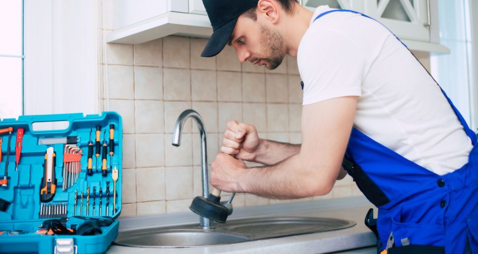 A plumber in a blue uniform and cap using a plunger in a stainless steel kitchen sink. A case with various tools is open on the counter.